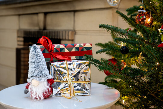 A cute Scandinavian Christmas gnome beside festive gifts wrapped in silver and geometric paper with a red bow on a white table by a fireplace and a glowing Christmas tree. A cozy holiday still life