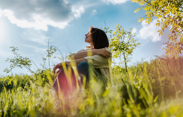 Obraz premium Low angle shot of a dreamy middle aged woman sitting on green grass against a beautiful sky with clouds