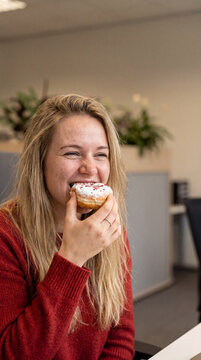 A young smiling blonde woman wearing a red sweater and taking a bite of a delicious powdered donut with colorful sprinkles