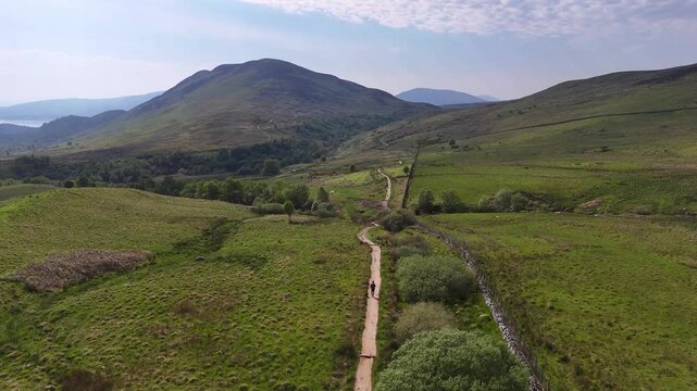 An elevated perspective glides over verdant, rolling terrain. A hiker walks along the winding trails of the West Highland Way on a bright spring afternoon towards Conic Hill in Scottland.
