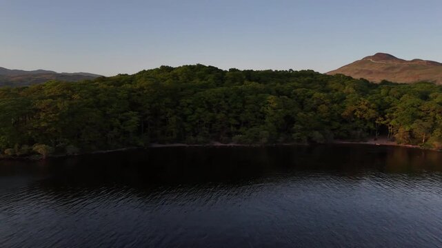Aerial drone shot of Loch Lomond along the West Highland Way in Scotland. The drone pans left over the lake showing the sandy shore and trees.