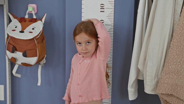 Girl places hand on head while measuring height against wall growth chart in building; growth milestone curiosity.