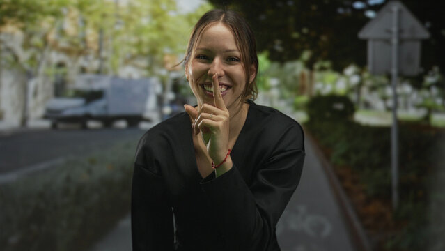 Young hispanic woman smiling, finger to lips for silence on a city street with bike lane and trees; playful secrecy.