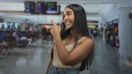 Fototapeta premium Hispanic woman points finger at departure board in airport, smiling broadly showing teeth; joyful anticipation.