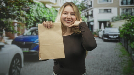 Fototapeta premium Woman smiling holds brown paper bag with hand and makes call gesture on residential street with parked cars; friendly delivery joy.
