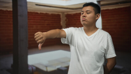 Fototapeta premium Man showing thumbs down with outstretched arm and visible thumb inside building interior with brick wall; disapproval critique.