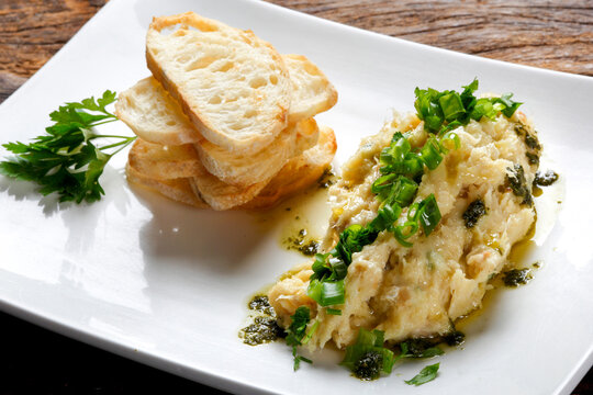 A delicious appetizer featuring shredded salted cod (bacalhau) drizzled with extra virgin olive oil and fresh green herbs, served with toasted crusty bread slices on a white plate.