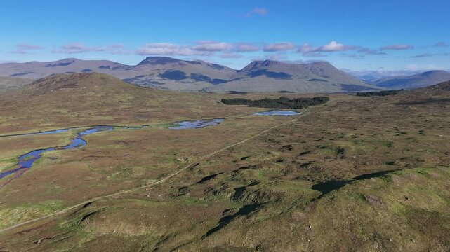 Aerial drone pan left and boom up shot of the West Highland Way in Scotland. It shows the empty Rannoch moor swamp area with small lakes and few trees. Mountains and blue sky in the back.
