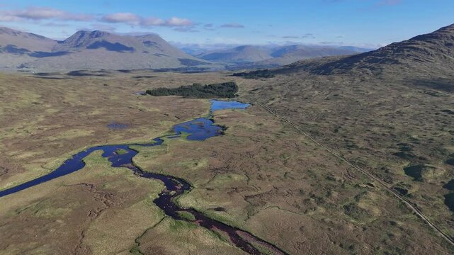 Aerial drone pan left and boom up shot of the West Highland Way in Scotland. It shows the empty Rannoch moor swamp area with small lakes and few trees. Mountains and blue sky in the back.
