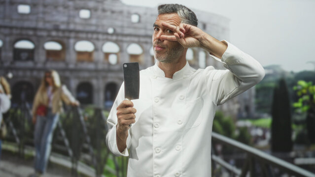 Grey haired man chef in white jacket holds cleaver in right hand and flashes v sign at eye in front of building colosseum; playful mischief.