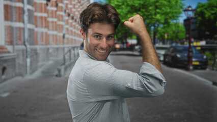 Fototapeta premium Young hispanic man smiles and flexes biceps while rolling up sleeve on a sunny urban street with parked cars and brick building facades; confidence playfulness.