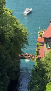 Wide shot of Ponte della Civera in Nesso, Lake Como, with crowds of young people gathering and jumping from the historic stone bridge into the turquoise water below. Summer adventure atmosphere.