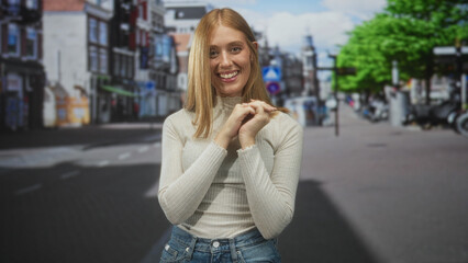 Fototapeta premium Redhead woman smiling and clasping hands near her face, wearing a turtleneck sweater and jeans while standing on a busy city street with storefronts; joy warmth.