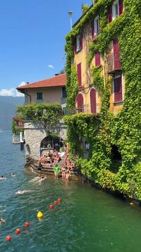 Nesso, Italy-05 30 2026: Young people swimming in the refreshing waters of Lake Como near a historic ivy-covered stone building Authentic summer lifestyle in a picturesque Italian village.