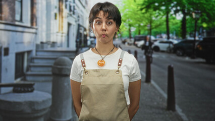 Obraz premium Woman wearing apron puckers lips, hands behind back on urban street with parked cars and stone bollards near building facade; curiosity exploration.