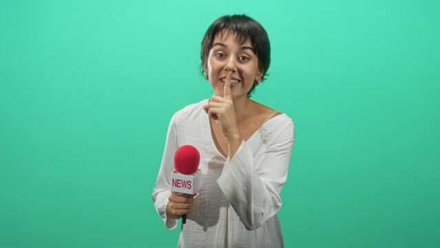 Woman reporter holding a news microphone and finger to lips for silence in a green studio, microphone flag shows news; secrecy hush.