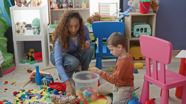 Woman kneeling, hand reaching for plastic blocks as young boy sorts toys on a playroom floor inside a building; learning calm.