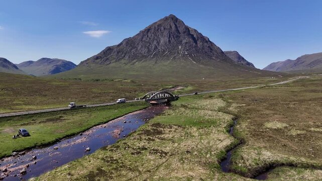 Aerial drone push forward and boom up shot of the Buachaille Etive Mor on the West Highland Way in Scotland. It captures the mountain with a round arch bridge and a car street in front of it.