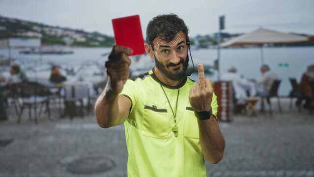 Referee wearing fluorescent shirt holds red card and shows middle finger on street by waterfront cafe, whistle and headset visible; defiance authority rebuke.