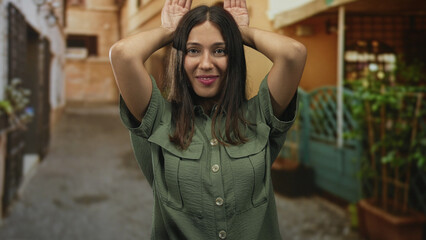 Fototapeta premium Hispanic brunette woman with hands on head posing on narrow street in green shirt, smiling with tongue out and playful expression; playful joy.