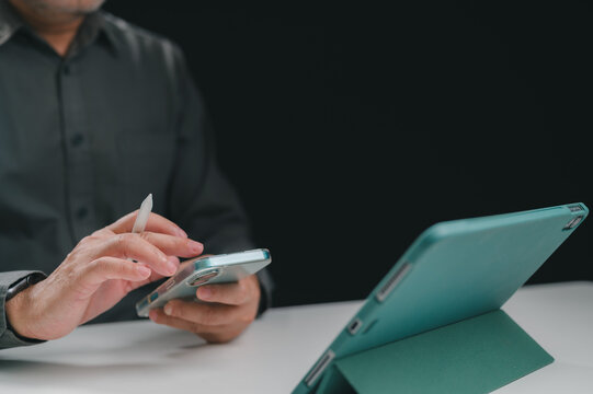 Mature male businessman wearing dark shirt holds stylus operating smartphone sitting desk opposite tablet computer inside dim office space late night working session multitasking digital tasks.