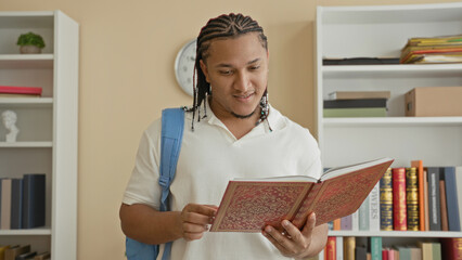 Man with hands holding an ornate book, backpack on shoulder, reading and turning pages in an indoor library room with bookshelves and wall clock  focused study. © Krakenimages.com