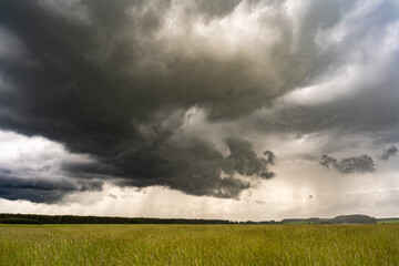 Obraz premium A massive dark storm cloud hangs low over a green meadow with rain visible in the distance