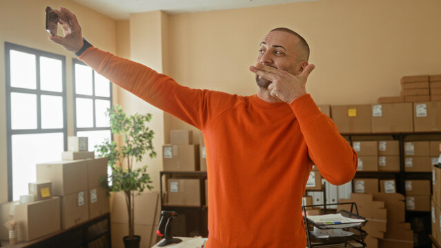 Man in orange sweater holding smartphone, pointing and posing among stacked boxes in a building parcel office; focused.