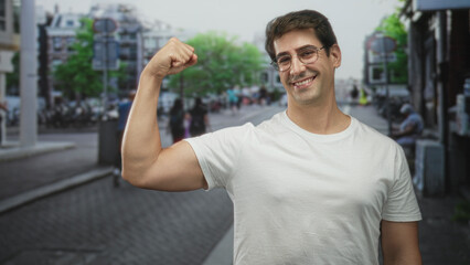 Fototapeta premium Man flexes bicep on city street while smiling and looking sideways in white t shirt; confidence success.