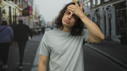 Fototapeta premium Man, young with long hair and glasses, hand on forehead in a busy city street wearing gray t shirt; fatigue reflection.