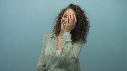 Fototapeta premium Woman laughing with hand covering face, wearing glasses and mint green polka dot shirt and curly hair in blue studio; joy authenticity warmth.