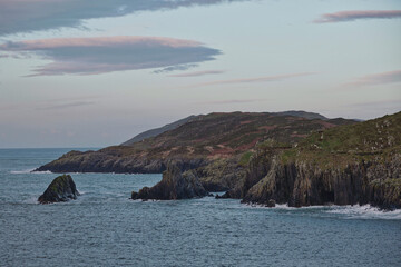 The ocean meets a rocky shoreline and sea stacks, with rolling hills covered in sparse vegetation forming the background. © Oleksii