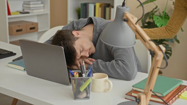 Man at laptop with woman touching his shoulder and leaning over while he glances up from a desk scattered with books and a lamp inside a building office; concern support.