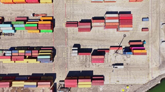 Cargo containers fill a shipping yard under clear skies. Trucks move around loading and unloading goods. The layout shows various colors and arrangements of containers.