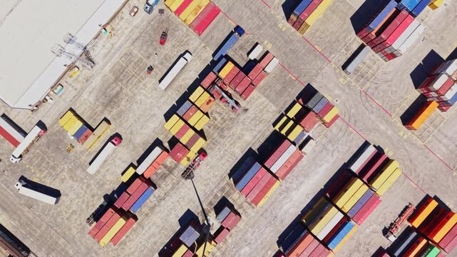 Workers move shipping containers at a large yard filled with colorful stacks. Trucks are parked nearby as equipment operates among rows of boxes.