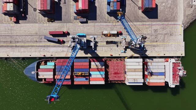 A cargo ship is at a port while containers are being loaded onto it. The area is active with cranes and many containers stacked around. It is daytime.