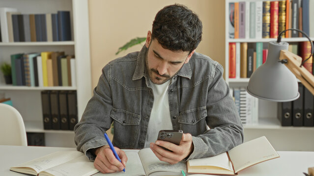 Man writing notes with pen while holding smartphone at a study desk in a building study room with bookshelves and desk lamp visible; concentration.