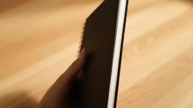 Womans Hand Holding a Black Notebook on a Wooden Surface.