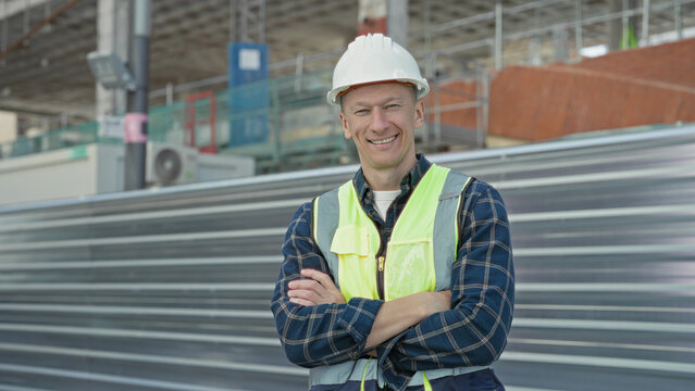 Man in hardhat and high visibility vest with arms crossed smiling at building site by corrugated metal fence and scaffolding; confidence.