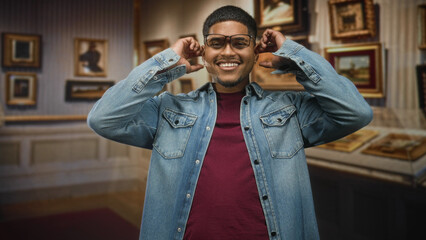Fototapeta premium Hispanic man adjusts glasses and smiles while touching ears amid framed paintings in an art gallery building; joy wonder.