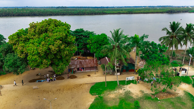Aerial view of a riverbank with lush green trees, small rustic buildings, and people walking along the sandy path near the water in Minima, Rivers, Nigeria.