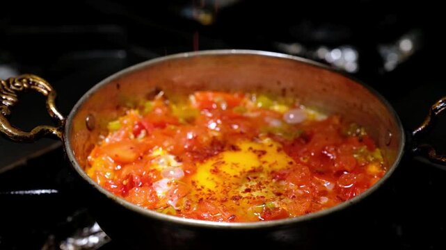 Tomatoes and green peppers cook over high heat in a copper pan as flames rise around the dish. Close-up scene showing menemen preparation, Turkish breakfast and restaurant kitchen cooking.