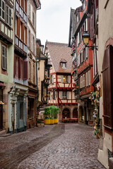 Obraz premium Intimate shuttered alley framed by red timbered facades, glossy cobbles, small planters and warm shopfronts, quiet rainy day mood ideal