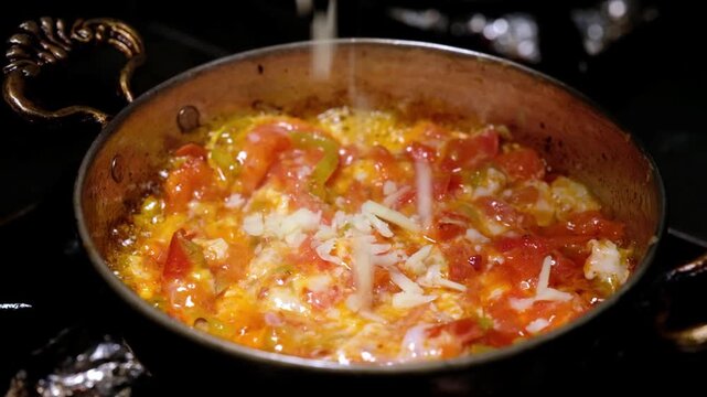 Tomatoes and green peppers cook over high heat in a copper pan as flames rise around the dish. Close-up scene showing menemen preparation, Turkish breakfast and restaurant kitchen cooking.
