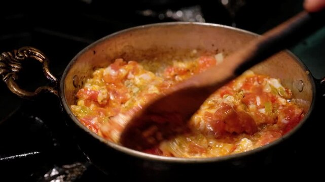 Tomatoes and green peppers cook over high heat in a copper pan as flames rise around the dish. Close-up scene showing menemen preparation, Turkish breakfast and restaurant kitchen cooking.