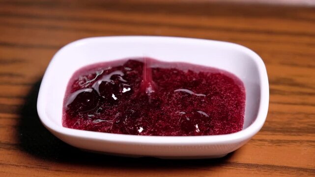 Red fruit jam is poured into a white serving bowl. Close-up scene showing breakfast jam presentation, a sweet breakfast spread and table serving detail.