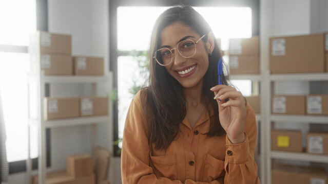 Young hispanic woman holding blue pen to cheek among stacked parcel boxes in small office building; thoughtful planning.