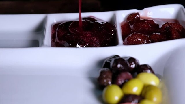 Red jam is served in a white serving plate with mixed olives placed nearby. Close-up scene showing breakfast jam service, sweet breakfast spread and table presentation detail.