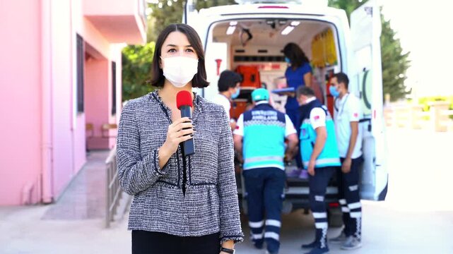 A woman wearing a face mask holds a microphone while medical staff transport a patient on a stretcher from an ambulance, conveying a sense of breaking news in the healthcare or emergency services fiel