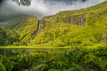 Impresionante vista panorámica de cascadas cayendo por un acantilado verde, reflejadas en un lago rodeado de densa vegetación, Flores, Azores. © Maxi Perez